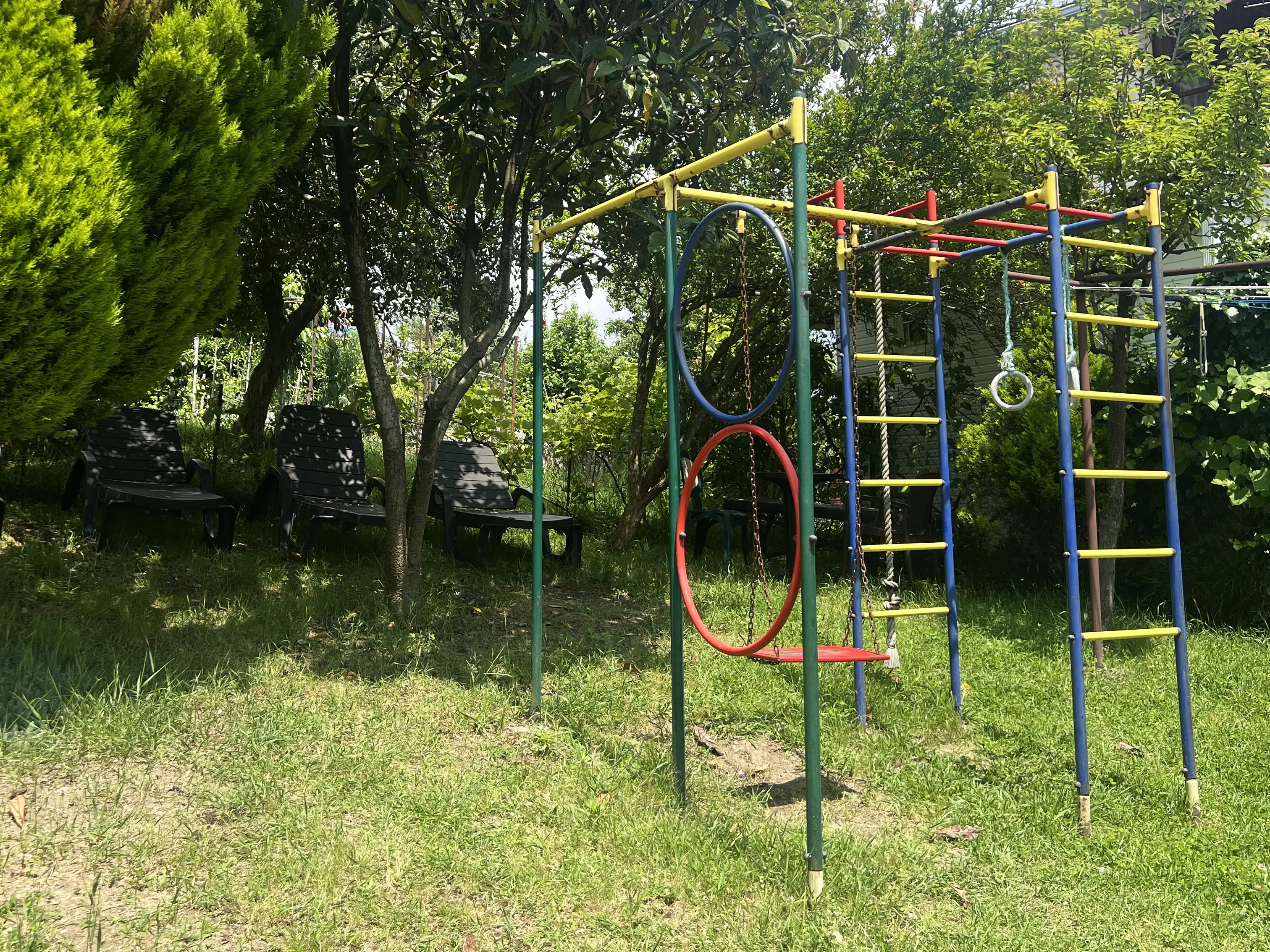 Детская площадка с качелями, лестницей и турником. Над площадкой дерево мушмулы слева и граната справа